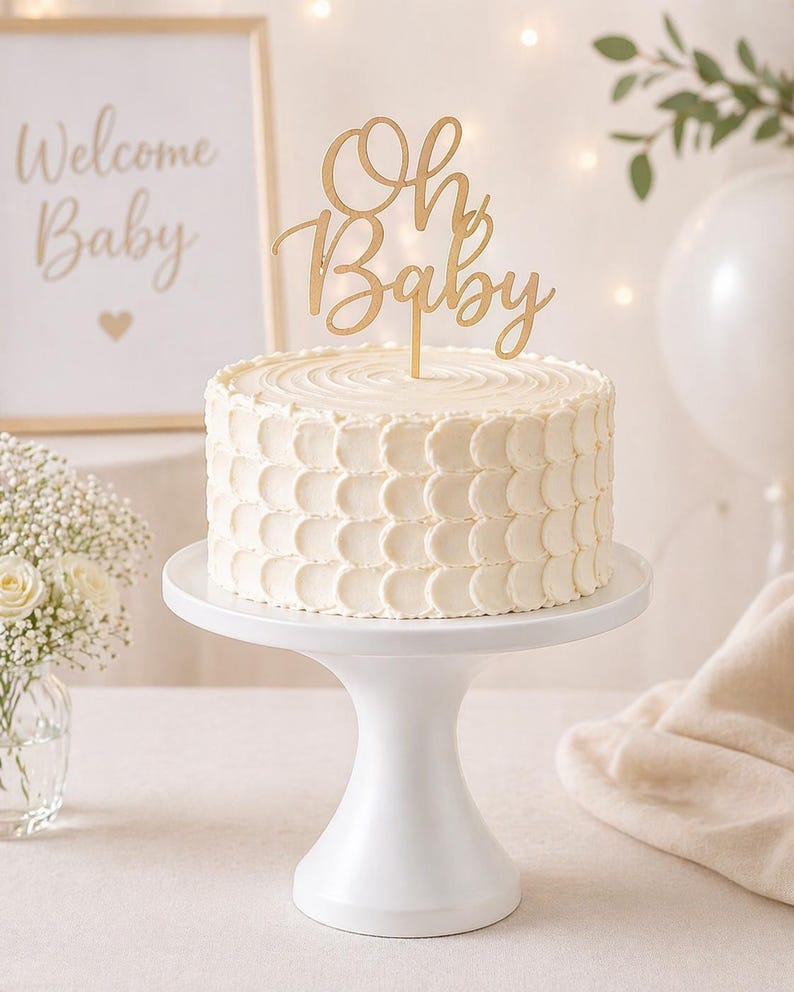 May include: A white frosted cake with a gold "Oh Baby" cake topper sits on a white cake stand. A framed sign that says "Welcome Baby" is in the background. The cake is decorated with a textured pattern.