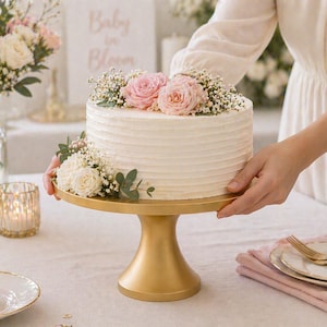 May include: A white frosted cake decorated with pink roses and greenery, presented on a gold cake stand. The cake is the centerpiece of a table setting, with a cupcake, flowers, and a candle in the background. The cake is for a baby shower.