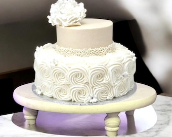 May include: A two-tiered white cake with buttercream frosting, decorated with piped rosettes and a pearl bead border. The top tier is topped with a large white flower. The cake sits on a round, light-colored cake stand.