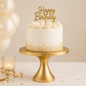 May include: A white frosted cake with gold sprinkles and a gold "Happy Birthday" cake topper sits on a gold cake stand. A cluster of cream-colored balloons and a lit candle in a glass holder are in the background.