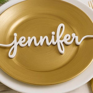 Könnte beinhalten: Ein weißes Namensschild mit dem Schriftzug "Jennifer" liegt auf einem goldfarbenen Teller, der sich auf einem weißen Teller befindet. Das Namensschild ist ein dekoratives Element, wahrscheinlich für eine Tischdekoration.