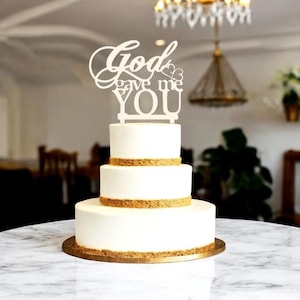 Può includere: Una torta bianca a tre strati con finiture dorate e un cake topper in legno con la scritta "God gave me YOU". La torta poggia su una base rotonda in legno ed è esposta su un tavolo di marmo.