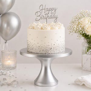 May include: A white frosted cake with silver sprinkles and a "Happy Birthday" cake topper. The cake sits on a silver cake stand. Silver balloons and a bouquet of white flowers are in the background.