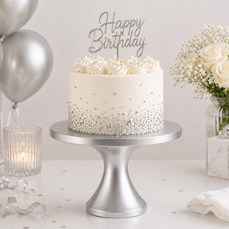 May include: A white frosted cake with silver sprinkles and a "Happy Birthday" cake topper. The cake sits on a silver cake stand. Silver balloons and a bouquet of white flowers are in the background.