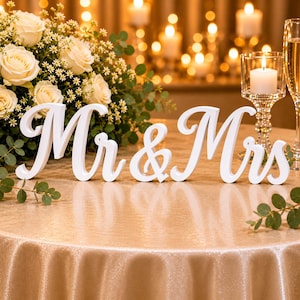 Op de afbeelding: Een wit houten bord met de cursieve tekst Mr & Mrs. Het bord staat op een tafel met een goudkleurig tafelkleed, vergezeld door een boeket witte rozen met groen blad, een glazen kaarsenhouder met een witte kaars en een champagneglas.