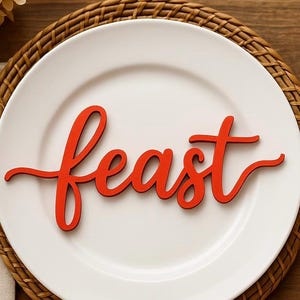 Puede incluir: Un plato blanco con la palabra "feast" en rojo, escrita en cursiva, sobre un salvamanteles tejido. Un tenedor y un cuchillo flanquean el plato, creando un arreglo de comedor. El entorno es sobre una mesa de madera.