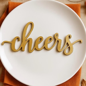 Może przedstawiać: Biały talerz ze złotym napisem "cheers" w centralnej części. Złoty widelec i nóż znajdują się po obu stronach talerza. Talerz leży na pomarańczowej serwetce, z małą dynią i jagodami w tle.