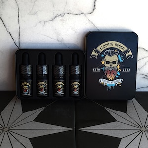 Könnte beinhalten: Ein Set aus vier kleinen schwarzen Flaschen mit goldenen Etiketten und Tropfern, beschriftet mit "Blueberry Pie", "Winter Splash", "Mystic Flannel" und "Grove Bay". Die Flaschen befinden sich in einer schwarzen Box mit einem Totenkopf-Logo und dem Text "Flaming Beard Est. 2023 Beard Oils and Candles".