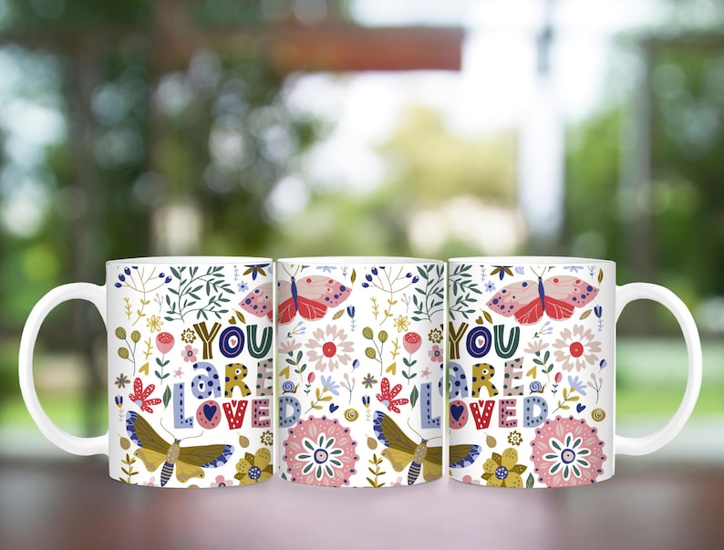 May include: Three white ceramic mugs with a floral pattern and the text "You are loved" in colorful letters. The mugs have a butterfly design and are perfect for a gift.