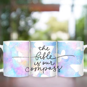 May include: Three white ceramic mugs with a watercolor-style background of blue, pink, and purple. The middle mug has the text "the bible is our compass" in black.