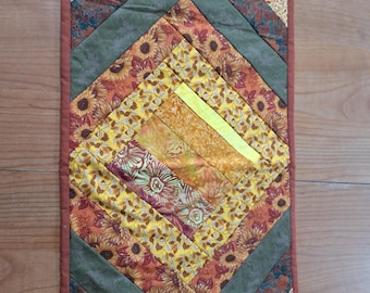 May include: A quilted table runner with a diamond pattern. The runner features various fabrics in shades of yellow, orange, green, and brown. The central diamond showcases sunflower and floral patterns, while the borders have leaf and solid-colored designs.