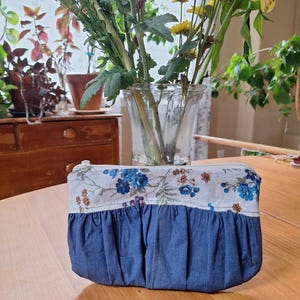 May include: A handmade denim and floral print zippered pouch. The top section features a white fabric with blue and brown floral patterns. The bottom is a gathered denim fabric. The pouch is sitting on a wooden table.