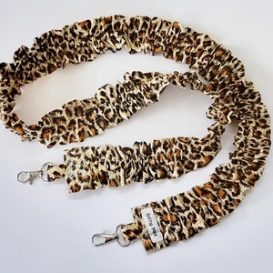 May include: A long, brown and tan leopard print camera strap with two silver clips. The strap is made of fabric and has a ruffled texture. The strap has a small white tag with the text "Dotty MK" on it.