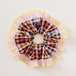 May include: A vibrant plaid scrunchie with a pink pleated ruffle and yellow edging. The scrunchie showcases a blend of pink, orange, black, yellow, and blue in a traditional plaid design. This hair accessory is designed for styling.