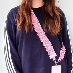 May include: A pink and white checkered phone lanyard with a ruffled design. The lanyard is attached to a pink phone case. The person in the image is wearing a navy blue sweatshirt with white stripes on the sleeves.