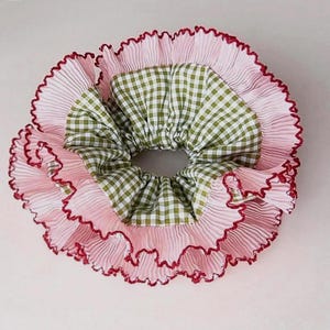 Könnte beinhalten: Ein rosa Haargummi mit gerüschtem Rand und einem grün-weiß karierten Mittelteil. Das Haargummi hat eine plissierte Textur und eine dunkelrote Bordüre, wodurch ein geschichteter Effekt entsteht. Dieses Haaraccessoire ist für verschiedene Frisuren geeignet.