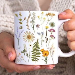 May include: White ceramic mug with a floral design featuring pressed flowers and ferns in shades of yellow, green, and purple.