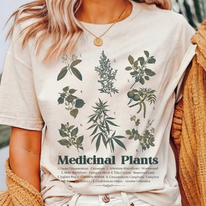 May include: A white t-shirt featuring a graphic print of various medicinal plants and their names. The text reads "Medicinal Plants" and lists the names of the plants, including Cinnamon, Wormwood, Basswood Lime, and Camphor.