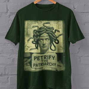 May include: Forest green t-shirt with a graphic of Medusa, her hair made of snakes, and the text "Petrify the Patriarchy." The design is in a rectangular frame. The shirt hangs on a wooden hanger against a grey brick wall.