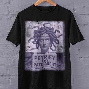 May include: Black t-shirt with a graphic of Medusa, depicted with snakes for hair, a white face, and the text "PETRIFY THE PATRIARCHY". The design is in purple and white. The t-shirt is displayed on a wooden hanger.