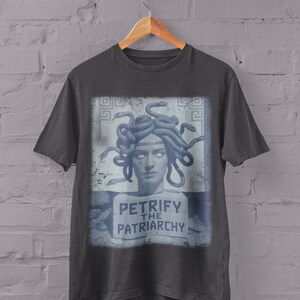 May include: Charcoal gray t-shirt featuring a graphic of Medusa with snakes for hair, and the text "PETRIFY THE PATRIARCHY" in a blue and white design. The shirt is hanging on a wooden hanger against a white brick wall.