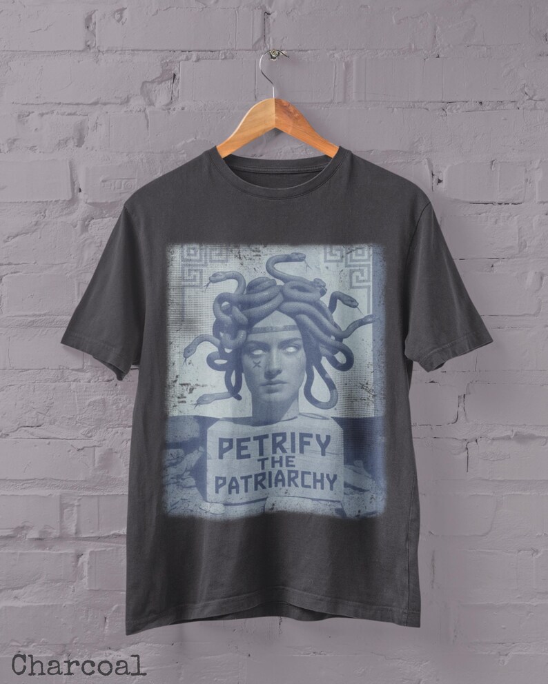 May include: Charcoal gray t-shirt featuring a graphic of Medusa with snakes for hair, and the text "PETRIFY THE PATRIARCHY" in a blue and white design. The shirt is hanging on a wooden hanger against a white brick wall.
