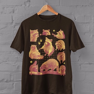 K&ouml;nnte beinhalten: Dunkel schokoladenfarbenes T-Shirt mit einer Collage von Waschb&auml;ren in verschiedenen Posen, mit einem gelb-rosa Farbschema. Das T-Shirt enth&auml;lt unten den Text "Dark Chocolate".