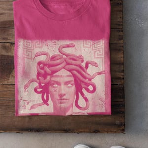 May include: A pink t-shirt featuring a Medusa graphic in a lighter pink hue. The design includes a portrait of Medusa with snakes for hair. The word "Heliconia" is printed at the bottom left.