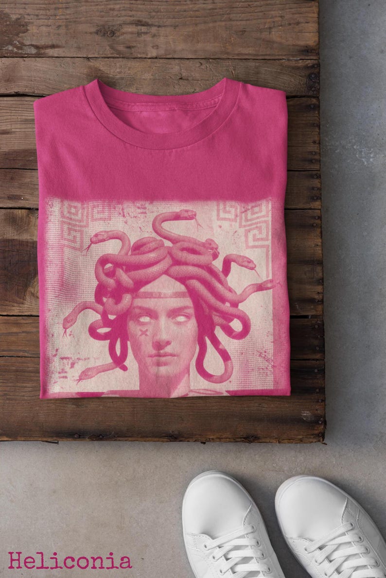 May include: A pink t-shirt featuring a Medusa graphic in a lighter pink hue. The design includes a portrait of Medusa with snakes for hair. The word "Heliconia" is printed at the bottom left.