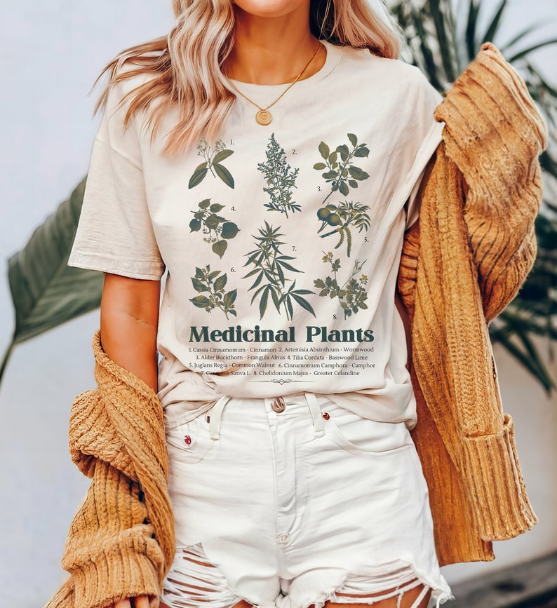 May include: A white t-shirt featuring a graphic print of various medicinal plants and their names. The text reads "Medicinal Plants" and lists the names of the plants, including Cinnamon, Wormwood, Basswood Lime, and Camphor.