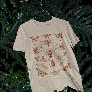 Pu&ograve; includere: T-shirt color sabbia con un motivo marrone di insetti e farfalle. La maglietta a maniche corte &egrave; appesa su uno sfondo di fogliame verde. La parola "Sand" &egrave; stampata in basso a sinistra.