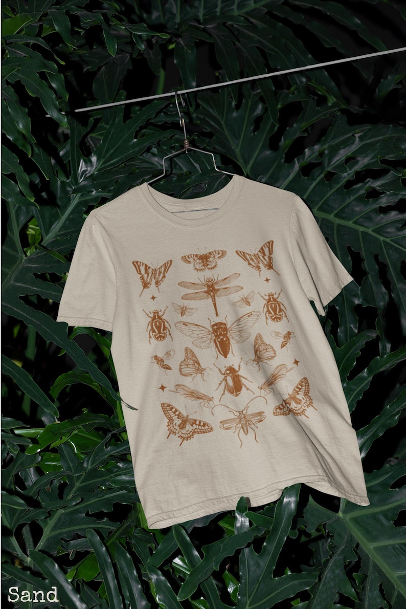 Pu&ograve; includere: T-shirt color sabbia con un motivo marrone di insetti e farfalle. La maglietta a maniche corte &egrave; appesa su uno sfondo di fogliame verde. La parola "Sand" &egrave; stampata in basso a sinistra.