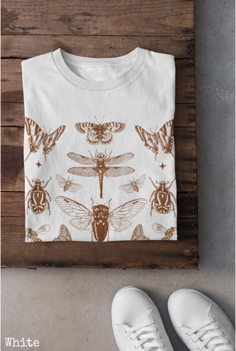 Pu&ograve; includere: T-shirt bianca con motivo a insetti marrone. Il design presenta illustrazioni dettagliate di farfalle, libellule, scarabei e altri insetti. La maglietta &egrave; piegata su una superficie di legno.