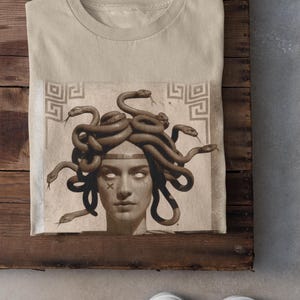 May include: Sand-colored t-shirt featuring a graphic of Medusa with snakes for hair. The artwork is in sepia tones, with a detailed depiction of the mythological figure. The shirt is folded on a wooden surface.