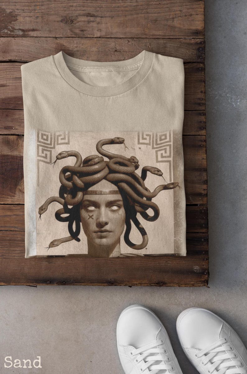 May include: Sand-colored t-shirt featuring a graphic of Medusa with snakes for hair. The artwork is in sepia tones, with a detailed depiction of the mythological figure. The shirt is folded on a wooden surface.