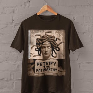 May include: Dark chocolate-colored t-shirt featuring a graphic of Medusa with snakes for hair. The image includes the text "PETRIFY THE PATRIARCHY" on a sign. The shirt is hanging on a wooden hanger against a brick wall.