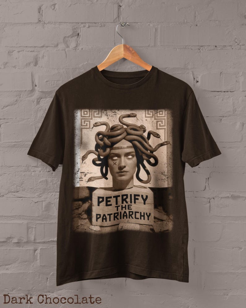 May include: Dark chocolate-colored t-shirt featuring a graphic of Medusa with snakes for hair. The image includes the text "PETRIFY THE PATRIARCHY" on a sign. The shirt is hanging on a wooden hanger against a brick wall.