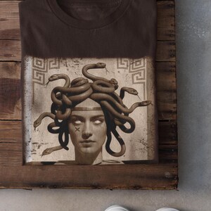 May include: Dark chocolate-colored t-shirt featuring a graphic of Medusa with snakes for hair. The image is printed on a rectangular patch with a vintage aesthetic. The shirt is folded on a wooden surface, with white sneakers visible.