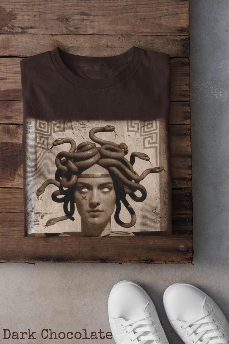 May include: Dark chocolate-colored t-shirt featuring a graphic of Medusa with snakes for hair. The image is printed on a rectangular patch with a vintage aesthetic. The shirt is folded on a wooden surface, with white sneakers visible.