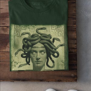 May include: Forest green t-shirt featuring a graphic of Medusa with snakes for hair. The image is set against a faded green background with Greek key patterns. The shirt is folded on a wooden surface, with white sneakers visible.