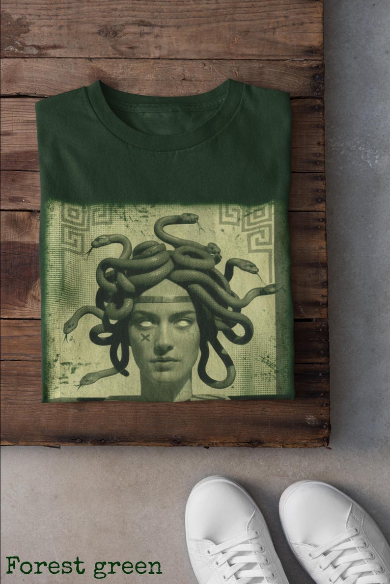 May include: Forest green t-shirt featuring a graphic of Medusa with snakes for hair. The image is set against a faded green background with Greek key patterns. The shirt is folded on a wooden surface, with white sneakers visible.