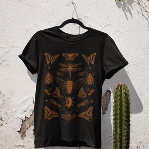 Pu&ograve; includere: T-shirt nera con un'illustrazione di insetti marroni. Il design include farfalle, coleotteri e libellule. La parola "Black" &egrave; stampata in basso a sinistra sulla maglietta.