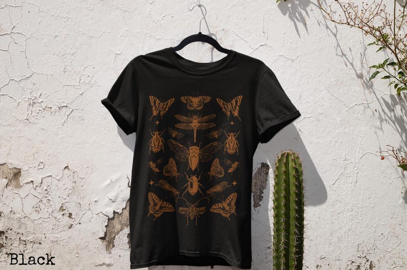 Pu&ograve; includere: T-shirt nera con un'illustrazione di insetti marroni. Il design include farfalle, coleotteri e libellule. La parola "Black" &egrave; stampata in basso a sinistra sulla maglietta.