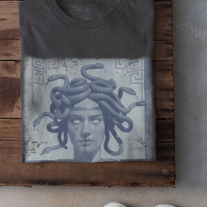 May include: Charcoal-colored t-shirt featuring a graphic of Medusa with snakes for hair. The design is in shades of blue and gray, with a distressed, vintage aesthetic. The shirt is folded on a wooden surface.