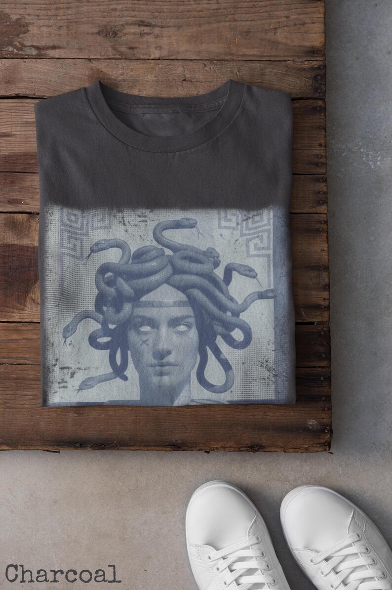 May include: Charcoal-colored t-shirt featuring a graphic of Medusa with snakes for hair. The design is in shades of blue and gray, with a distressed, vintage aesthetic. The shirt is folded on a wooden surface.