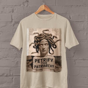 May include: Sand-colored t-shirt featuring a graphic of Medusa with snakes for hair. The image includes the text "PETRIFY THE PATRIARCHY" on a sign. The shirt is hanging on a wooden hanger against a brick wall.