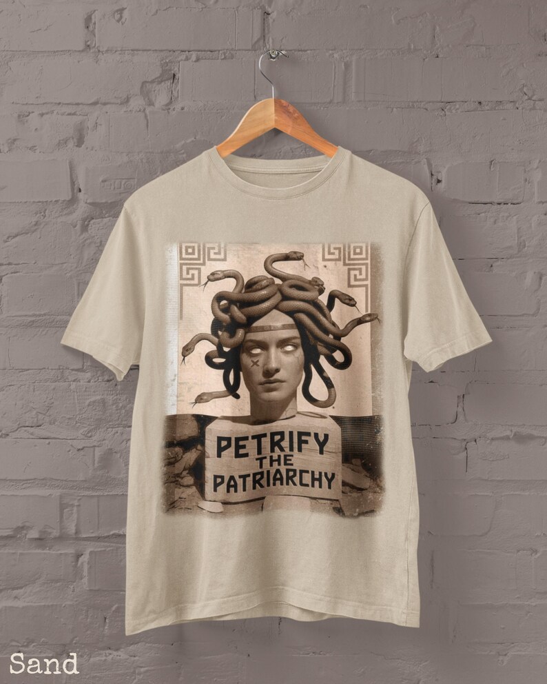 May include: Sand-colored t-shirt featuring a graphic of Medusa with snakes for hair. The image includes the text "PETRIFY THE PATRIARCHY" on a sign. The shirt is hanging on a wooden hanger against a brick wall.
