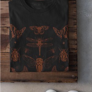 Pu&ograve; includere: T-shirt nera con un disegno di insetti marroni. Il disegno comprende farfalle, una libellula, scarabei e cicale. La parola "Black" &egrave; stampata in basso a sinistra dell'immagine.