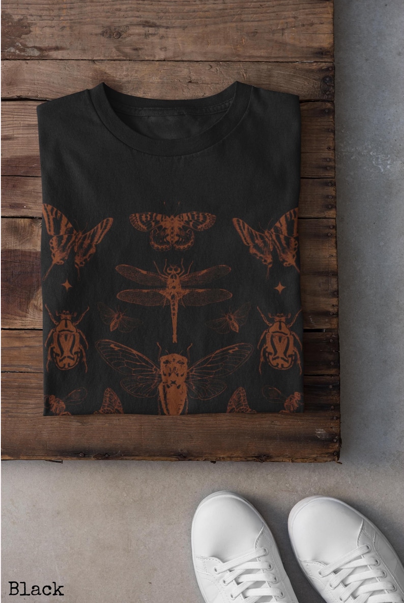 Pu&ograve; includere: T-shirt nera con un disegno di insetti marroni. Il disegno comprende farfalle, una libellula, scarabei e cicale. La parola "Black" &egrave; stampata in basso a sinistra dell'immagine.