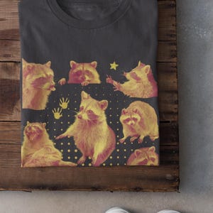 K&ouml;nnte beinhalten: Anthrazitfarbenes T-Shirt mit einem Design aus gelben und rosa Waschb&auml;ren in verschiedenen Posen. Das Design umfasst einen Stern und Handabdr&uuml;cke auf dunklem Hintergrund. Das Wort &bdquo;Charcoal&ldquo; ist unten aufgedruckt.